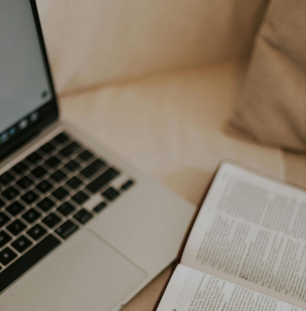An open Bible next to a laptop on a cozy bedspread, ideal for studying or devotional moments.