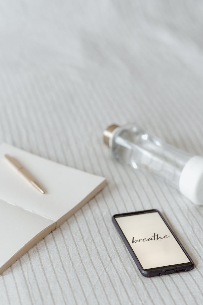 From above of cellphone with Breathe inscription on screen near small bottle of water and empty prayer notebook with pen