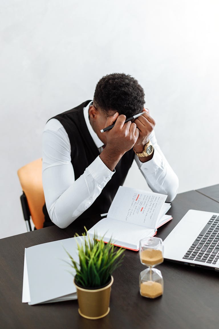 A businessman overwhelmed working indoors with a notebook, laptop, and hourglass.