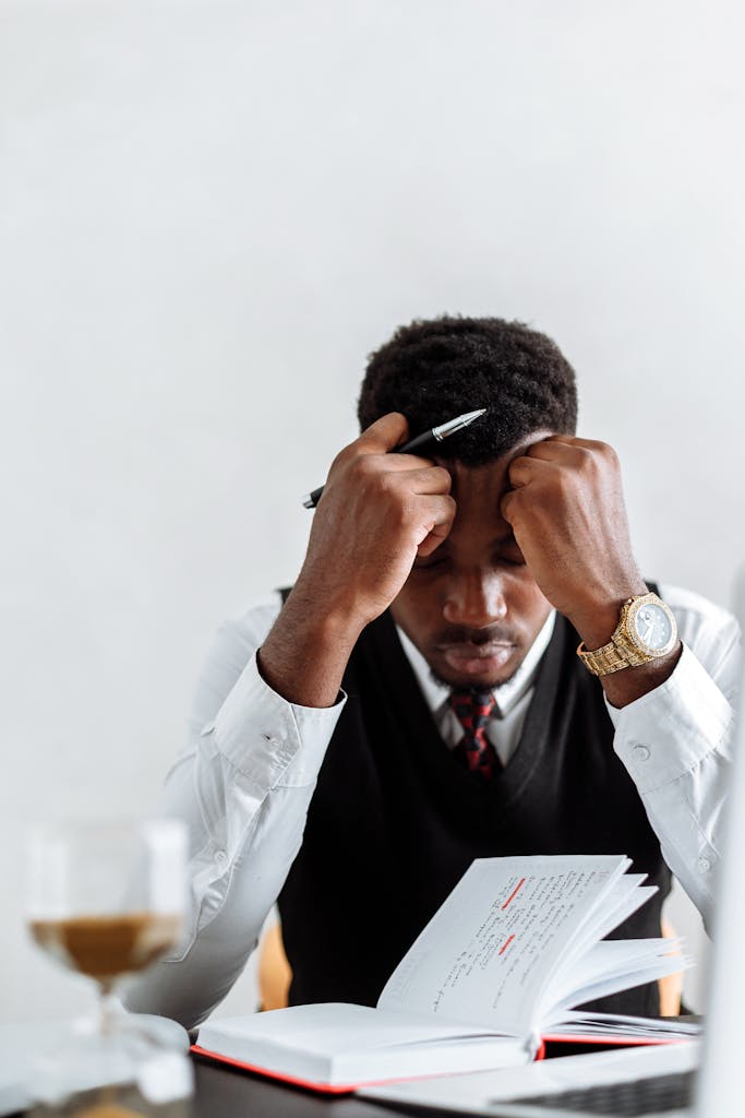 An overwhelmed businessman sits at a desk reviewing documents, conveying stress and concentration, possibly praying.