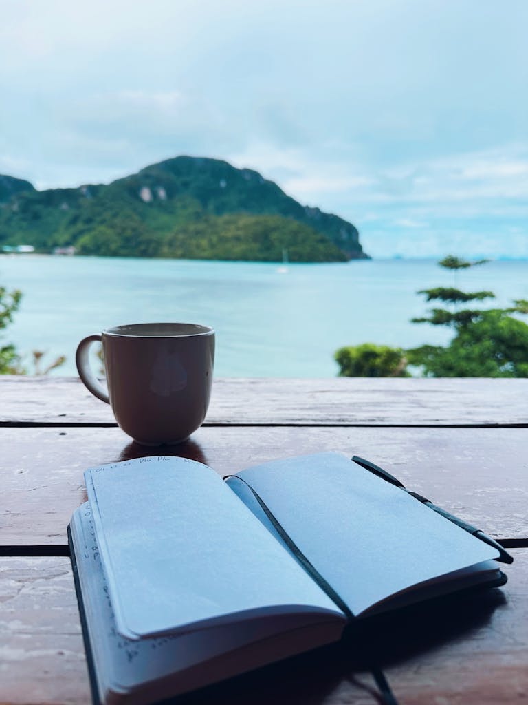 Open prayer journal and coffee cup on a table by a serene beachside view.