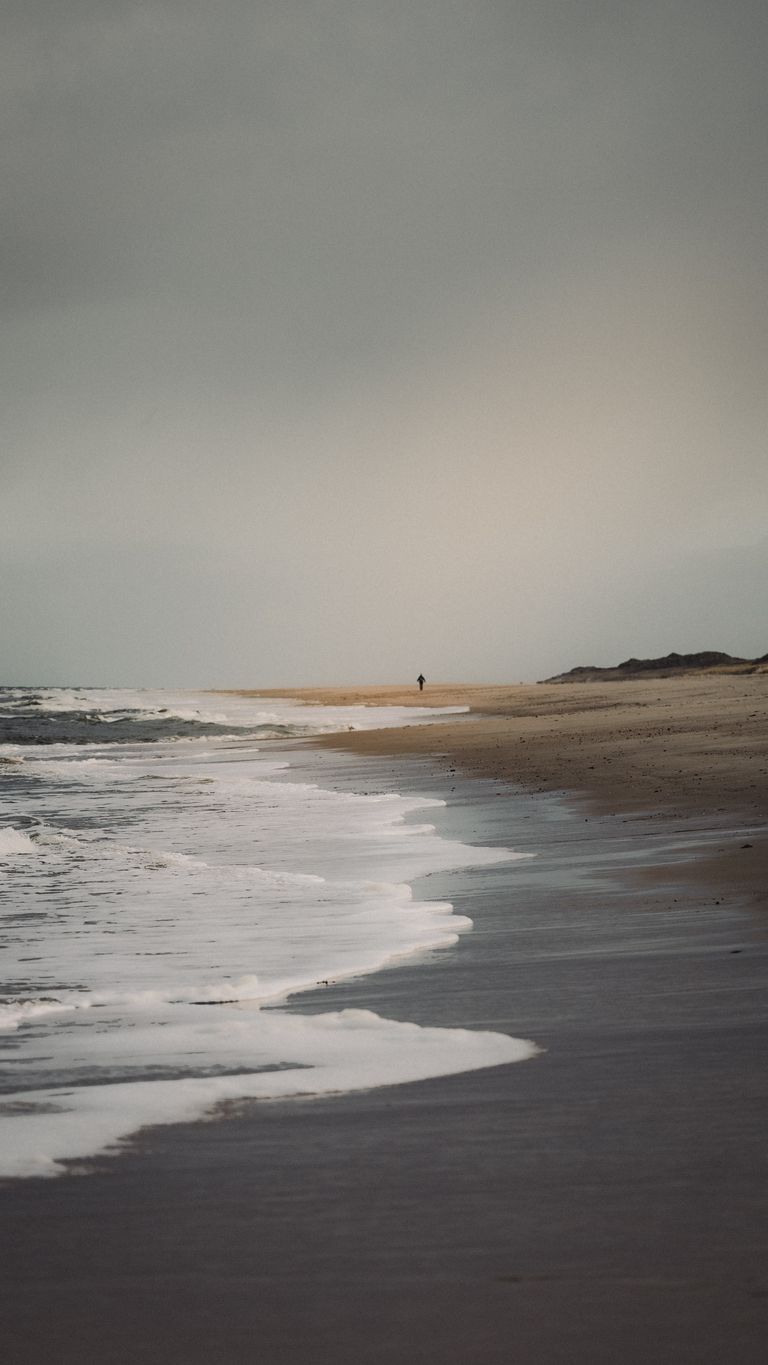 A solitary figure walks along a tranquil beach under a cloudy sky, capturing a sense of peace and solitude.