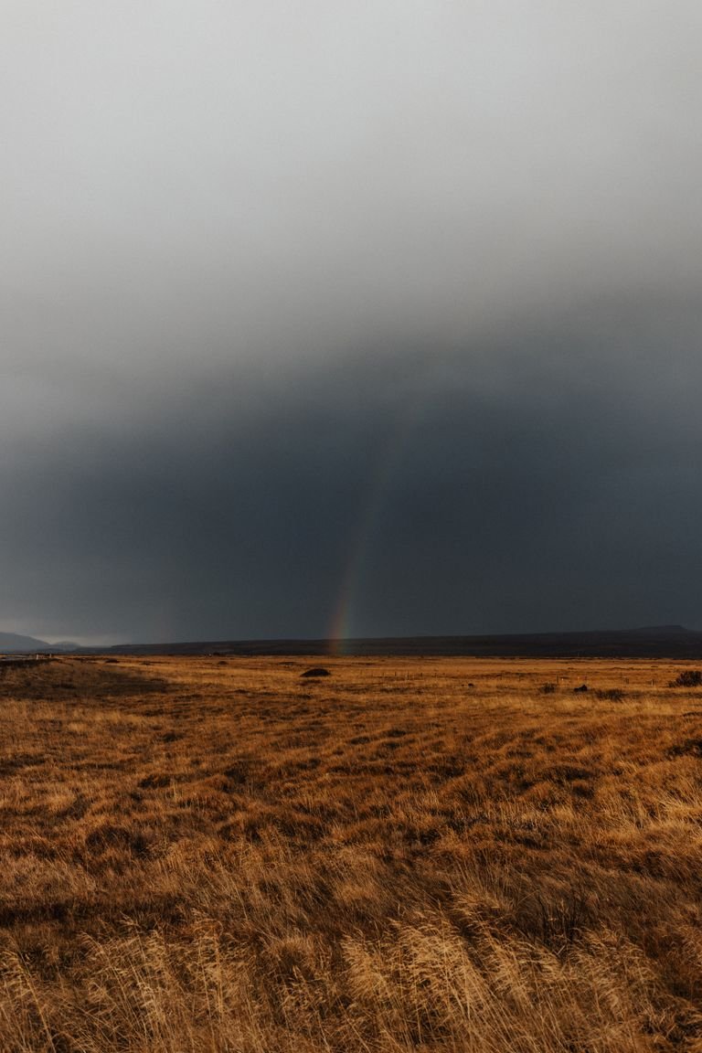 Moody countryside scene with fields and a subtle rainbow under an overcast sky.