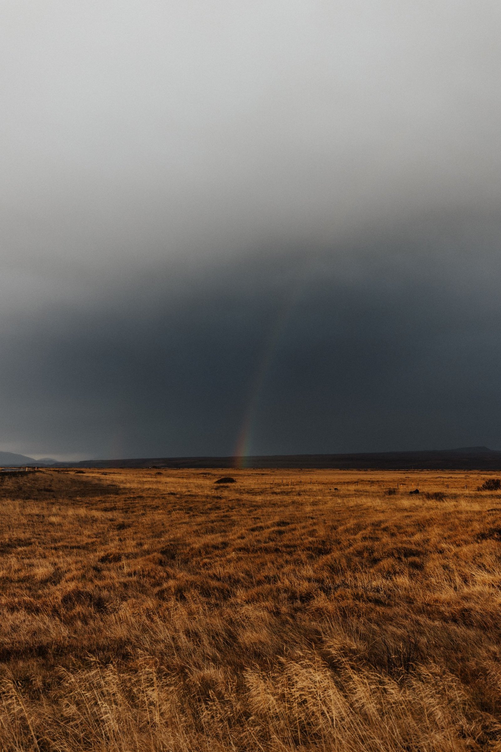 Moody countryside scene with fields and a subtle rainbow under an overcast sky.