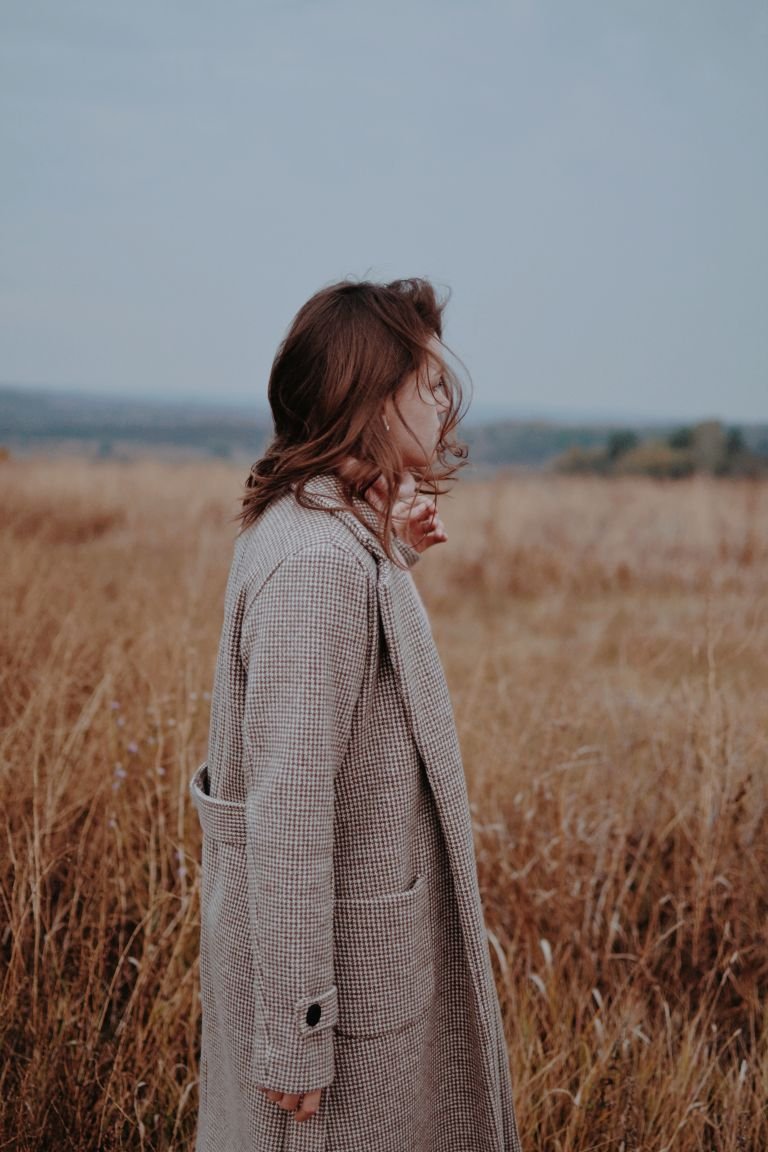 A brunette woman in a checkered coat stands looking at a serene grass field.