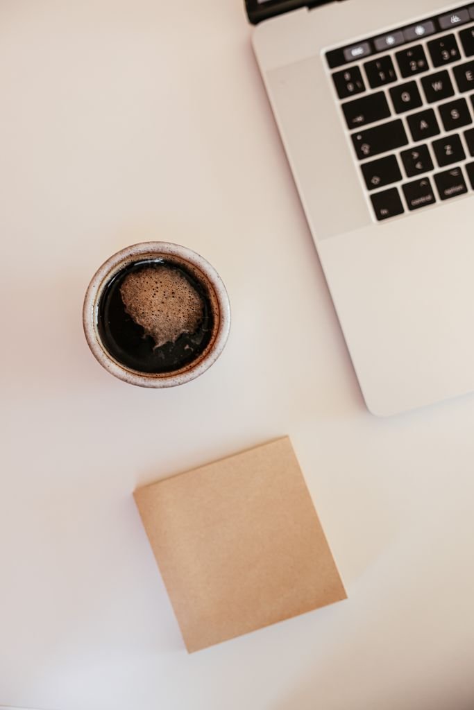 A minimalist workspace featuring a cup of coffee, laptop, and notepad on a white desk.