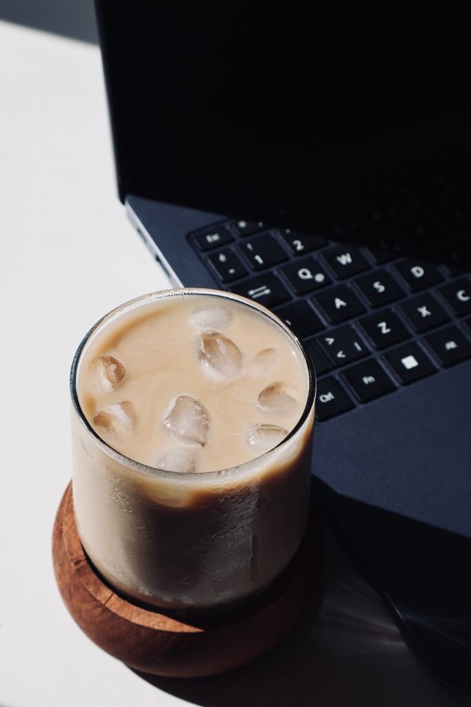 Biblical Mindset & Behavioral Coaching :A refreshing glass of iced coffee milk beside a laptop in bright sunlight, perfect for a work break.