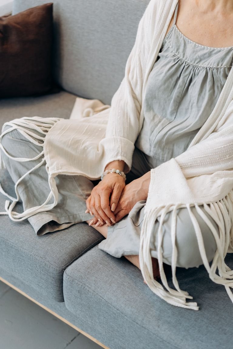 An elderly woman sits cross-legged on a sofa, hands together.